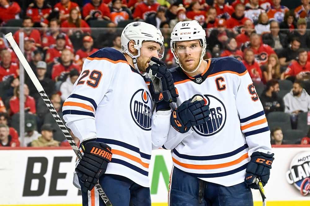 Edmonton Oilers forwards Leon Draisaitl and Connor McDavid stand on ice in white jerseys talking before taking a face-off.
